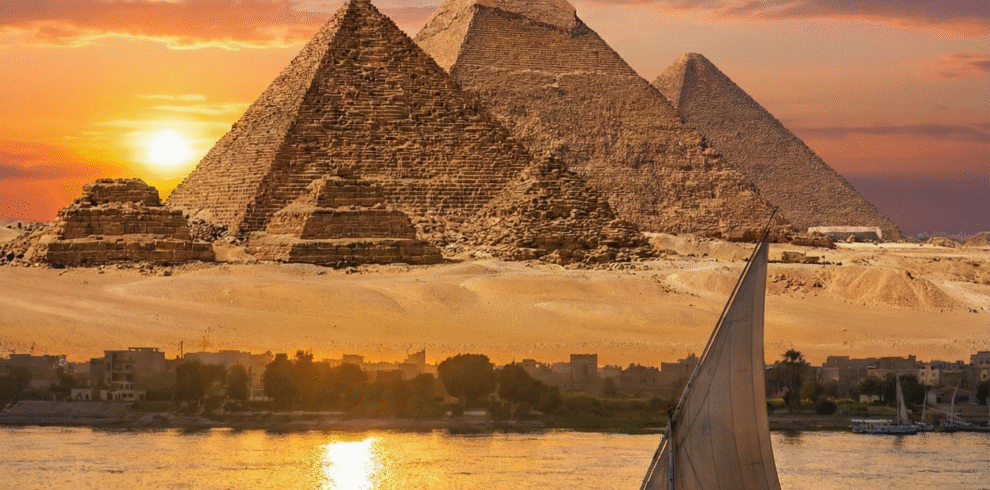 Panoramic view of the Great Pyramids of Giza at sunset, with a traditional felucca sailboat sailing on the Nile River. A classic scene for Cairo sightseeing tours and excursions.