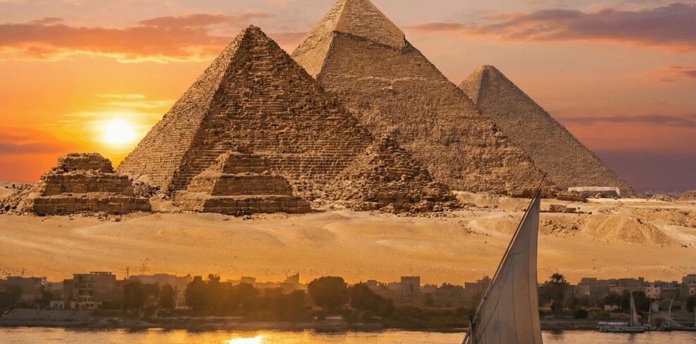 Panoramic view of the Great Pyramids of Giza at sunset, with a traditional felucca sailboat sailing on the Nile River. A classic scene for Cairo sightseeing tours and excursions.
