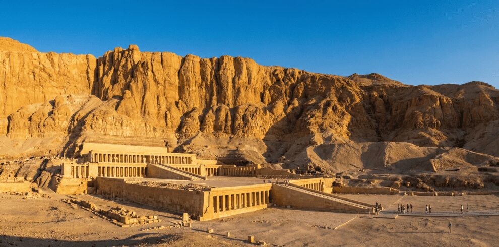 Wide cinematic shot of the Mortuary Temple of Hatshepsut in Luxor, majestic limestone terraces against a clear blue sky and steep cliffs, tourists walking in the distance, high resolution, sunny day, golden hour lighting.